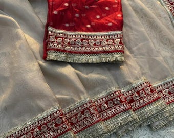 May include: A beige sheer fabric with a red accent piece. The red accent has intricate silver embroidery and a fringe trim. The fabric is draped, creating soft folds and shadows. The overall aesthetic is elegant and detailed.