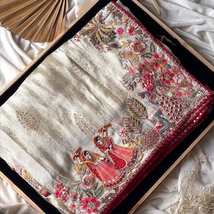 May include: An ivory-colored sari with intricate embroidery, featuring floral and peacock motifs in red, pink, and green. The sari has a striped pattern and a red border. The design includes two figures in traditional attire.
