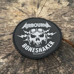 May include: A black and white embroidered patch featuring the text "AIRBOURNE" and "BONESHAKER" in a stylised font. The patch depicts a skull wearing a helmet with lightning bolts. The text "RNRFL" and "©2019 AIRBOURNE" are also included.