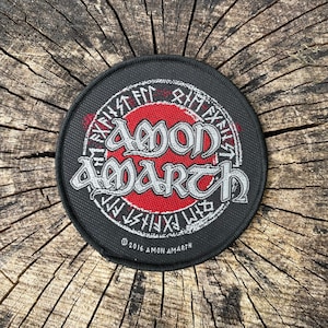 Puede incluir: Un parche con el logotipo de la banda Amon Amarth con un círculo rojo y letras blancas. El parche es negro con un círculo blanco alrededor del logotipo y runas alrededor del borde exterior. El texto "© 2016 AMON AMARTH" está en la parte inferior del parche.