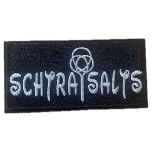 May include: A rectangular black patch with white embroidered text that reads "SCHYRA SALTS". Above the word "SALTS" is a white embroidered design. The patch is likely for clothing or accessories.