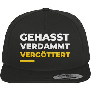 Könnte beinhalten: Schwarze Snapback-Kappe mit den deutschen Wörtern "GEHASST VERDAMMT VERGÖTTERT" in Weiß und Gelb. Die Kappe hat einen flachen Schirm und einen Schnappverschluss auf der Rückseite.