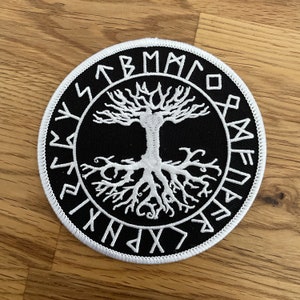 Puede incluir: Un parche bordado en blanco y negro con un diseño de árbol de la vida con raíces y ramas. El árbol está rodeado por un círculo de runas blancas.