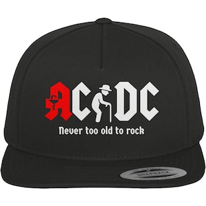 Op de afbeelding: Zwarte snapback pet met de tekst "AC/DC" in een gestileerd lettertype, een rood logo en een witte afbeelding van een persoon met een wandelstok. De slogan "Never too old to rock" staat eronder.
