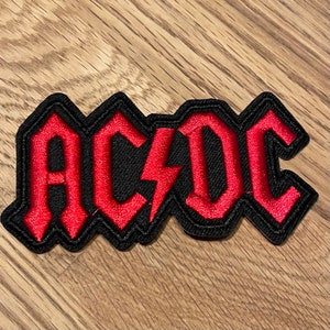 May include: A red embroidered AC/DC logo patch with a black background. The logo is in a bold, stylized font with a lightning bolt in the centre.