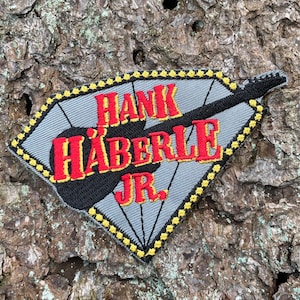 May include: A gray diamond-shaped patch with a black guitar and the text "Hank Häberle Jr." in red and yellow lettering.