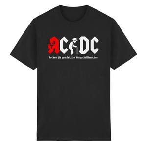 Könnte beinhalten: Schwarzes T-Shirt mit einem weißen und roten Grafikdesign. Das Design enthält den Text "AC/DC" in einer stilisierten Schriftart, mit dem Bild einer Person mit einem Stock. Darunter steht der Text "Rocken bis zum letzten Herzschrittmacher".