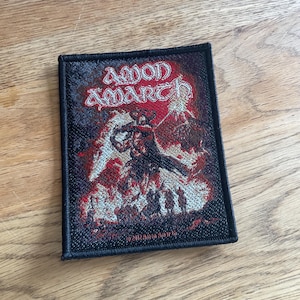 May include: A black embroidered patch featuring the band name "Amon Amarth" in white lettering. The patch depicts a red and white image of a horned figure wielding a sword, surrounded by flames and a stormy sky.