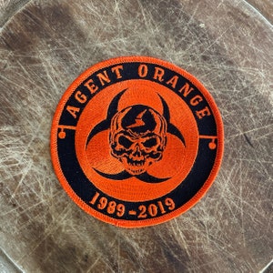 May include: A black and orange embroidered patch with a skull and biohazard symbol. The text "AGENT ORANGE 1989-2019" is stitched around the edge.