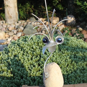 Könnte beinhalten: Eine skurrile Metallvogel-Skulptur mit einem Steinkörper, die auf einer Holzkiste steht. Der Vogel hat glänzende Metallaugen, einen Schnabel und eine Krone aus Metallstäben. Die Skulptur steht vor einem Hintergrund aus grünem Laub.