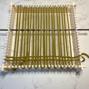 May include: A white plastic loom with a wooden dowel and a mustard yellow cord woven through it.
