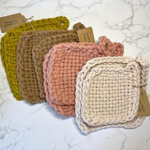 Puede incluir: Conjunto de cuatro agarraderas de olla de crochet hechas a mano en amarillo, marrón, rosa y crema. Las agarraderas de olla están hechas con un patrón tejido y tienen una forma cuadrada.