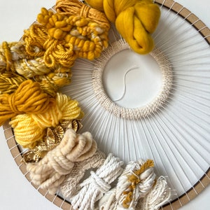 May include: Circular woven wall hanging with a natural wood frame. The design features a sunburst pattern with white string radiating from a central woven circle. Accents include various shades of yellow and cream yarn, creating a textured, artistic piece.