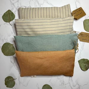 May include: Four rectangular linen eye pillows in different colours: beige with black stripes, light blue, and orange. The pillows are stacked on top of each other on a white marble surface. Each pillow has a small tag attached with handwritten text.
