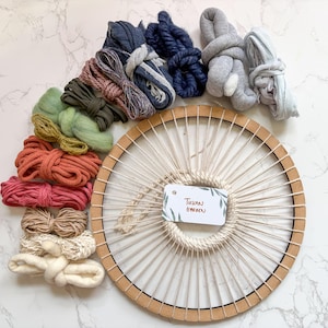 Könnte beinhalten: Ein Bastelset mit einem kreisförmigen Holzrahmen und verschiedenen Bündeln farbigen Garns in Blau-, Grün-, Rosa- und Beigetönen. Ein Etikett mit dem Text "Tuscan Garden" ist am Rahmen befestigt. Das Garn ist um den Rahmen angeordnet.