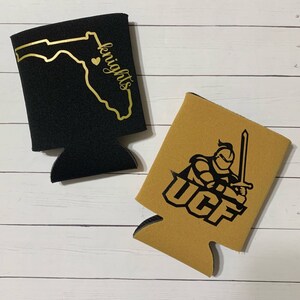 Op de afbeelding: Twee zwarte en gouden blikjeskoelers. Een koeler heeft een zwarte omtrek van de staat Florida met een hart en het woord "knights". De andere koeler heeft een zwarte ridder die een zwaard vasthoudt met de letters "UCF" in zwart.