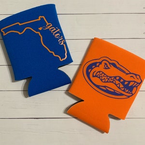 Op de afbeelding: Twee schuimrubberen blikjeskoelers. E&eacute;n is blauw met een oranje omtrek van de staat Florida en de tekst "gators". De andere is oranje met een blauwe alligator-kopgrafiek.