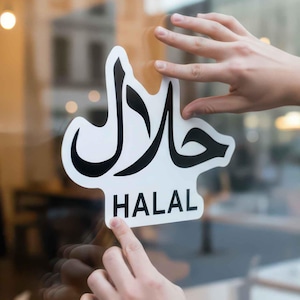 Op de afbeelding: Een wit en zwart "HALAL" bord wordt op een glazen raam aangebracht. Het bord bevat Arabisch schrift boven het Engelse woord "HALAL". De achtergrond toont een vage straatscène met gebouwen en mensen.