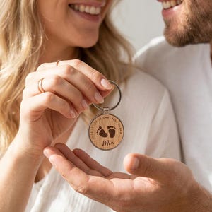 K&ouml;nnte beinhalten: Ein runder Holzschl&uuml;sselanh&auml;nger mit silbernem Metallrand und Ring. Das Holz zeigt eine Gravur von zwei Babyfu&szlig;abdr&uuml;cken, dem Namen Mia und dem Text 10.04.21, 09:15, UMK, 2680 GRAMM, die kreisf&ouml;rmig um die Mitte angeordnet sind.