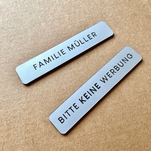 May include: Two rectangular silver-colored metal signs with rounded corners. The top sign features the black text FAMILIE MÜLLER, and the bottom sign features the black text BITTE KEINE WERBUNG. Both signs have a smooth, matte finish.