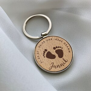 K&ouml;nnte beinhalten: Ein runder Holzschl&uuml;sselanh&auml;nger mit einem silbernen Metallring. Die Holzoberfl&auml;che ist mit zwei Babyfu&szlig;abdr&uuml;cken, dem Namen Jannik, dem Datum 21.05.21, der Uhrzeit 06:30 UHR und dem Gewicht 4345 GRAMM in dunkelbrauner Gravur versehen.