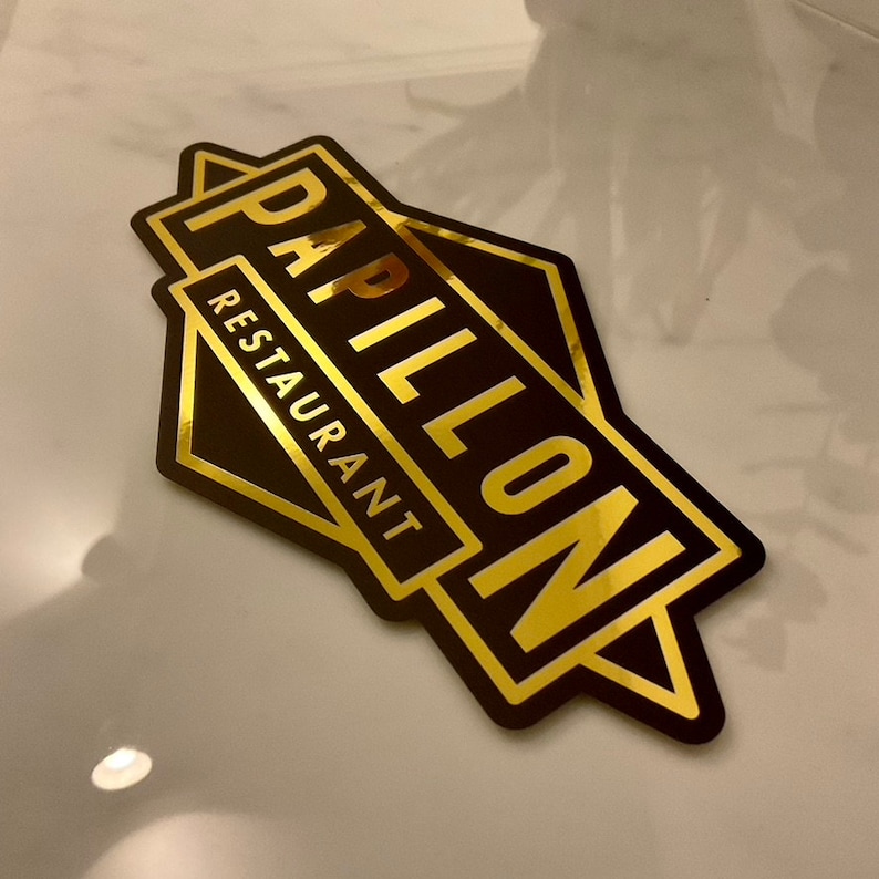 May include: A black and gold sticker with the words "Papillon Restaurant" in a stylized font. The sticker has a geometric design with gold lettering and accents, and is likely used for branding or advertising.