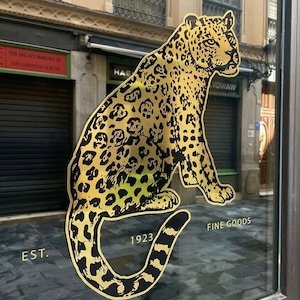 May include: A gold and black leopard graphic is displayed on a glass storefront. The leopard is sitting with its tail curled. The words "EST. 1923 FINE GOODS" are printed below the leopard.