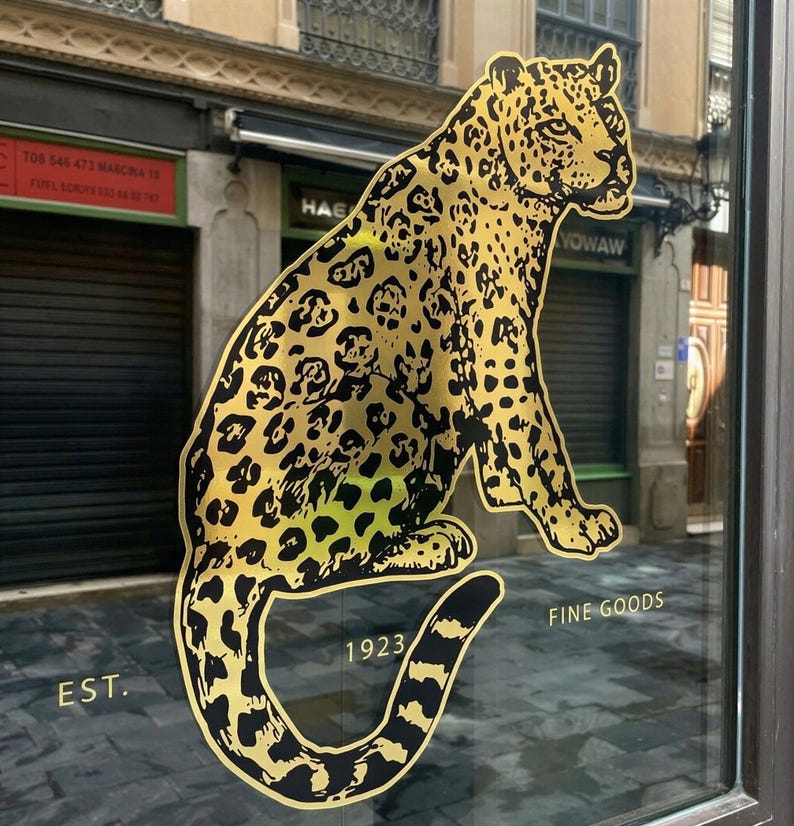 May include: A gold and black leopard graphic is displayed on a glass storefront. The leopard is sitting with its tail curled. The words "EST. 1923 FINE GOODS" are printed below the leopard.