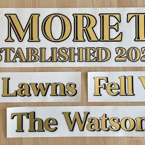 May include: Several white rectangular decals with gold lettering and black outlines. The largest decal reads "ONE MORE TIME ESTABLISHED 2022." Smaller decals say "The Lawns," "Fell View," and "The Watson."