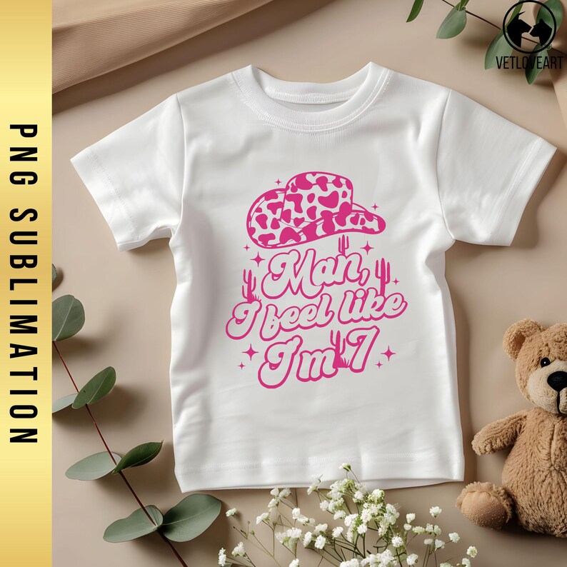 May include: White t-shirt with a pink cow print hat, cactus, and the text "Man, I feel like I'm 7".