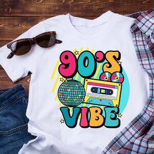 May include: White t-shirt with a colorful graphic design featuring the text "90's Vibe" with a disco ball, cassette tape, and stars.