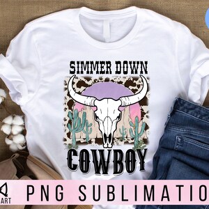 May include: White t-shirt with a graphic design of a cow skull with a pink sunset behind it. The text "Simmer Down Cowboy" is printed below the skull.