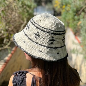 May include: A woman wearing a black crochet top with a white crocheted bucket hat. The hat has a black trim and a geometric pattern.