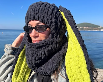 May include: A dark gray knitted balaclava with long, bright green and black bunny ears. The balaclava covers the head and neck, with a face opening. The ears are a striking contrast against the blue sky and water background.