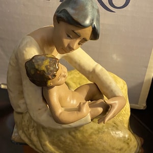 Può includere: Una statuetta in porcellana di una donna che tiene un bambino. La donna indossa un vestito giallo e ha i capelli castani. Il bambino è nudo e ha i capelli castani. La statuetta è realizzata da Lladró.