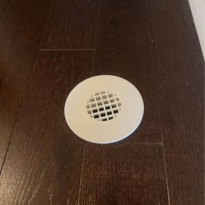 Op de afbeelding: Een ronde, witte afvoerkap met een zwart rooster in het midden, geplaatst in een donkerbruine houten vloer.