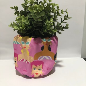 May include: A pink fabric pot cover decorated with various Barbie doll faces wearing sunglasses and gold earrings. The pot cover holds a green leafy plant. The word "Barbie" is printed on the fabric.