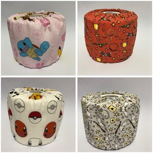 Puede incluir: Cuatro fundas de rollo de papel higiénico de tela. Una es rosa con un patrón de Squirtle y Pikachu. Otra es roja con un patrón de trofeo y gafas doradas. La tercera es crema con un patrón de Pokemon. La última es gris con un patrón de pájaro volador.