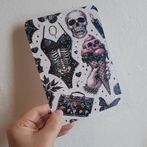 May include: A holographic sticker sheet with a black and white design featuring a skull, a skeleton in a swimsuit, an ice cream cone, a butterfly, and a small suitcase. The background is white with a glittery effect.
