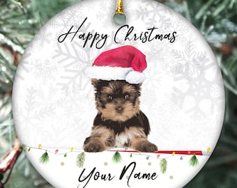 May include: A round Christmas ornament with a white background and a snowflake pattern. A Yorkshire Terrier puppy wearing a Santa hat is sitting on a red and white striped surface. The ornament says "Happy Christmas" and "Your Name".