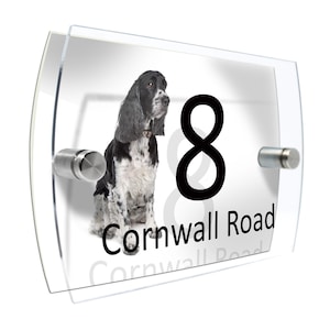Peut inclure: Panneau de numéro de maison en acrylique transparent avec une illustration de chien noir et blanc. Le numéro "8" est affiché en noir, avec le nom de la rue "Cornwall Road". Le panneau est monté avec des entretoises argentées.