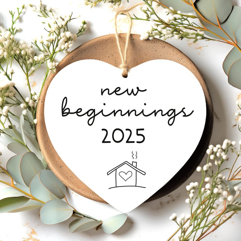 May include: White heart-shaped ornament with the text "new beginnings 2025" and a simple house drawing with a heart inside. The ornament is hanging from a string and is surrounded by white flowers.