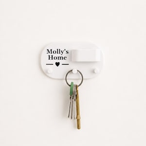 Könnte beinhalten: Ein weißer, ovaler Wand-Schlüsselhalter mit drei kleinen Haken und einem rechteckigen Schlitz. Der Text "Molly's Home" ist in Schwarz aufgedruckt, ergänzt durch ein kleines schwarzes Herzsymbol. Ein Metallschlüsselring mit mehreren Schlüsseln hängt am mittleren Haken.