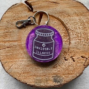 May include: Purple resin keychain with a white outline of a pill bottle that says "Invisible Illness".