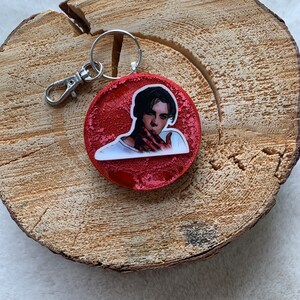 May include: A red, circular keyring with a silver clasp and ring. It features a graphic of a person with dark hair and a white shirt. The red background has a glittery texture.