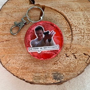 May include: Red round keychain featuring a picture of a man holding a gun. The text "We prefer the term psychotic" is in a speech bubble.