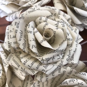 Puede incluir: Primer plano de varias rosas de papel hechas con p&aacute;ginas de libros. Los p&eacute;talos est&aacute;n cuidadosamente elaborados, mostrando el texto impreso. Las rosas son de color crema, con el texto en negro. El tema general es una artesan&iacute;a literaria y art&iacute;stica.