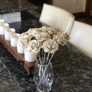 Puede incluir: Un ramo de rosas de papel hechas con p&aacute;ginas de libros en un jarr&oacute;n de cristal transparente. Las rosas son de color beige y blanco, con tallos de alambre plateado. El jarr&oacute;n est&aacute; sobre una encimera de granito oscuro. Velas blancas en el fondo.