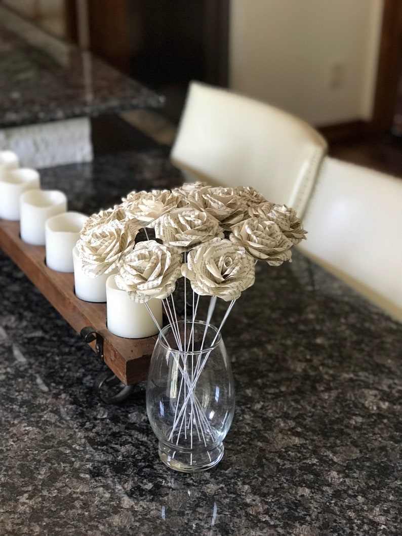 Puede incluir: Un ramo de rosas de papel hechas con p&aacute;ginas de libros en un jarr&oacute;n de cristal transparente. Las rosas son de color beige y blanco, con tallos de alambre plateado. El jarr&oacute;n est&aacute; sobre una encimera de granito oscuro. Velas blancas en el fondo.