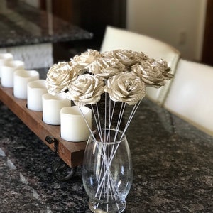Puede incluir: Un jarr&oacute;n de cristal transparente contiene un ramo de rosas de papel. Las rosas est&aacute;n hechas de p&aacute;ginas de libro, con tallos de alambre blanco. Un portavelas de madera con velas blancas est&aacute; a la izquierda sobre una encimera de granito oscuro.